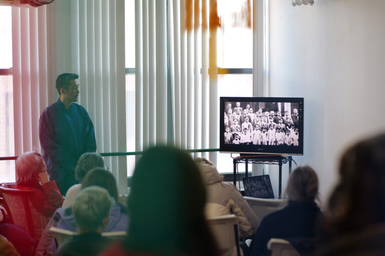 Photo of a man standing in front of a crowd. Everyone is watching a film on a TV.